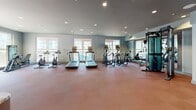 This fitness center is a comprehensively outfitted facility designed to support a wide range of workout routines. The space is laid out across a single floor, with durable rubber flooring throughout that provides both safety and functionality for high-intensity training.

The cardio area is generously sized, accommodating multiple treadmills, elliptical machines, and stationary cycling bikes. Large windows along the walls allow abundant natural light to fill the space, creating an open and energizing atmosphere for exercise. A cable machine and additional strength training stations are positioned nearby, offering versatility for resistance-based workouts.

A dedicated functional training zone features an extensive rack system stocked with a full range of kettlebells, medicine balls, and resistance bands, making it ideal for dynamic and circuit-style training. A separate strength area houses a multi-functional power rack with weight plates and a bench press setup for more traditional lifting.

The space is finished with a neutral blue-grey color palette on the walls, complemented by recessed lighting that ensures consistent illumination throughout. Exposed ductwork adds an industrial accent near the ceiling in certain areas, while glass-panel entry doors contribute to the modern aesthetic. The overall layout is open and well-organized, facilitating ease of movement between equipment zones.
