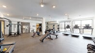 Located at 10 Commerce Dr in New Rochelle, NY, this fitness facility offers a well-appointed, single-floor layout designed to support a full range of exercise and wellness activities. The main gym area features rubber-floored training zones equipped with cardio machines including treadmills, ellipticals, and stationary bikes, as well as a cable machine, adjustable benches, and a rack of free weights. Large floor-to-ceiling windows along the perimeter allow natural light to fill the space while providing views of the surrounding exterior. Ceiling fans and recessed lighting throughout ensure a comfortable and well-lit environment during any workout. Adjacent to the main gym is a dedicated studio room finished with light hardwood flooring and a full-length wall mirror, making it ideal for yoga, stretching, or personal training sessions. A separate media room, also with hardwood flooring, features a wall-mounted smart TV, offering a versatile space for cooldown, recovery, or group screenings. The clean, neutral color palette and modern finishes give the facility a polished, professional aesthetic, while the thoughtful layout ensures ample space for movement and a variety of training modalities.