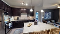 This single-floor space is designed with an open-concept layout that promotes a natural flow between the kitchen and living areas. The kitchen is a standout feature, showcasing rich dark espresso cabinetry that extends to the upper walls, providing ample storage. A mosaic tile backsplash adds texture and visual interest behind the sink, while granite countertops in a light speckled finish offer generous workspace. A curved kitchen island anchors the space and doubles as a casual dining area, illuminated by elegant pendant lights that hang from above. Track lighting supplements the overhead illumination, ensuring the kitchen is well-lit throughout. Stainless steel appliances, including a dishwasher, are seamlessly integrated into the cabinetry for a cohesive look. The open sightline from the kitchen into the living area reveals a comfortable lounge space accented with blue-toned artwork and warm furnishings, lending the overall interior a contemporary yet inviting ambiance. Light tile flooring throughout ties the spaces together while keeping the atmosphere bright and airy. The thoughtful design and quality finishes make this a functional and stylish single-floor residence.