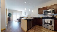 This well-designed single-floor residence in Fitchburg, Wisconsin, offers a thoughtfully arranged open-concept layout that seamlessly connects the kitchen, dining, and living areas. The kitchen is a standout feature, outfitted with rich dark cabinetry, dark granite countertops, and a full suite of stainless steel appliances including a refrigerator, range, built-in microwave, and dishwasher. A peninsula with bar seating provides additional workspace and casual dining, while three stylish pendant lights hang overhead, adding a contemporary touch. Warm hardwood-style flooring flows throughout the main living areas, enhancing the cohesive aesthetic. The living room is generously proportioned, anchored by a large sliding glass door with vertical blinds that admits abundant natural light and provides outdoor access. The bedroom offers a comfortable, serene retreat with a ceiling fan, natural light from a window with blinds, and ample closet access. The bathroom is finished with a large vanity featuring dark cabinetry, a wide mirror, a well-lit vanity bar, and a tub/shower combination. Throughout the residence, neutral wall tones and clean architectural lines create a bright, modern atmosphere that balances style with everyday functionality.