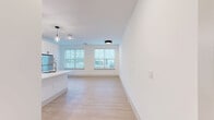 Located at 5025 Huntly Ridge St in Charlottesville, VA, this one-bedroom, one-bathroom unit offers a thoughtfully designed open-concept layout filled with natural light. Light-toned luxury vinyl plank flooring flows throughout the main living area, giving the space a clean, contemporary feel. The kitchen serves as a focal point, featuring a large central island with an undermount sink, crisp white cabinetry, stone countertops, and a full suite of stainless steel appliances including a refrigerator, range, and microwave. Stylish pendant lights hang above the island, adding a modern touch. The open living area extends from the kitchen, illuminated by multiple windows fitted with blinds that offer both light control and privacy. The spacious bedroom is carpeted for comfort and includes a ceiling fan, a generously sized window with natural light, and access to a dedicated walk-in closet. The bathroom is finished with a wide vanity, white quartz countertop, black-framed mirror, and matching black hardware accents, maintaining the unit's modern aesthetic. Clean white walls and recessed lighting throughout create a bright, inviting ambiance ideal for comfortable everyday living.