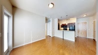 This single-floor space offers a thoughtfully designed open-concept layout that seamlessly connects the living area to the kitchen. The living space is bright and airy, featuring warm hardwood flooring that extends throughout, clean white walls, and a flush-mount ceiling light that provides even illumination across the room. A hallway leads to additional areas of the unit, with a closet visible nearby, adding practical storage to the layout.

The kitchen is a standout feature, defined by rich dark wood cabinetry that provides ample storage and a sleek granite countertop peninsula. The peninsula serves as both a functional workspace and a casual dining or entertaining surface, anchored by a bold black support post. Black appliances, including a microwave and refrigerator, complement the dark cabinetry and contribute to a cohesive, modern aesthetic. Overhead, a set of pendant lights and track lighting illuminate the kitchen workspace with focused, warm light, creating a welcoming atmosphere.

The open flow between the living and kitchen areas makes the space feel expansive and well-suited for both everyday living and entertaining. The neutral color palette, quality finishes, and warm wood flooring combine to deliver a comfortable and stylish interior throughout.