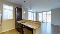 This contemporary single-floor residence showcases a thoughtfully designed open-concept layout that seamlessly connects the kitchen and living areas. The kitchen is a standout feature, boasting sleek dark cabinetry that provides a striking contrast against light granite countertops. A peninsula-style island with an undermount sink serves as both a functional workspace and a natural dividing point between the kitchen and the adjoining living space. Stainless steel appliances, including a dishwasher, complement the modern aesthetic, while pendant lighting with vintage-style fixtures adds a warm, inviting ambiance overhead. The living area benefits from an abundance of natural light, courtesy of large floor-to-ceiling windows that frame views of the surrounding streetscape. Light hardwood flooring throughout the space enhances the airy, open feel. The neutral wall tones and clean architectural lines create a versatile backdrop suited to a variety of interior styles. The overall design balances modern functionality with understated elegance, making this a bright and welcoming space ideal for both everyday living and entertaining.