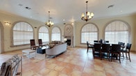 This single-floor space showcases a refined Mediterranean-inspired aesthetic that combines classical architectural details with an open, airy layout. The main living and dining area is defined by a series of grand arched windows dressed with white plantation shutters, allowing abundant natural light to fill the room throughout the day. Warm terracotta tile flooring extends across the open-plan space, lending a timeless, earthy character to the interior. A striking stone fireplace serves as a central focal point, flanked by ornate carved detailing and decorative column accents that reinforce the classical design language. Crown molding runs along the perimeter of the ceiling, adding a polished, architectural finish overhead. Two elegant chandelier fixtures hang from the ceiling, providing warm ambient lighting that complements the soft, neutral color palette of cream and beige tones. The living area is furnished with a classic rolled-arm sofa and accent chairs arranged around a central coffee table, while the adjacent dining area features a dark wood table with high-back chairs. Together, these spaces create a cohesive, inviting environment well-suited for both everyday living and formal entertaining.