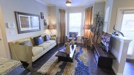 This single-floor living space showcases a thoughtfully designed interior with a cohesive and sophisticated aesthetic. The living room serves as the focal point, featuring rich dark hardwood floors that contrast elegantly with the neutral-toned walls accented by classic crown molding. A large bay window arrangement floods the space with natural light, complemented by floor-length gold and bronze drapery that adds warmth and texture to the room.

Seating is anchored by a cream-colored sofa adorned with deep blue and navy accent pillows, paired with a coordinating blue armchair positioned near the window. A dark wood coffee table sits atop a multi-toned shag area rug in blue, cream, and brown tones, tying the color palette together cohesively. Decorative figurines and tabletop accents add visual interest throughout.

Ambient lighting is provided by two matching slender table lamps with warm-toned shades, while a ceiling-mounted light fixture reinforces the room's classic character. A media console along one wall and a tall indoor plant add functional and natural elements. Overall, the space exudes a warm, polished ambiance suited for comfortable yet stylish everyday living.