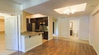 This single-floor residence offers a thoughtfully designed open-concept layout that seamlessly connects the kitchen and living areas. Rich hardwood flooring flows throughout the main living space, adding warmth and continuity to the design. The kitchen is a standout feature, equipped with stainless steel appliances including a refrigerator and built-in microwave, complemented by dark espresso cabinetry that provides a striking contrast against the light-toned walls. A granite-topped breakfast bar with seating overhang serves as both a functional workspace and a casual dining area. Mosaic tile backsplash adds texture and visual interest behind the cooktop. The open passageway leads into a bright hallway, illuminated by a classic multi-bulb chandelier that adds an elegant touch to the space. Natural light filters through the recessed ceiling areas, enhancing the airy atmosphere. A full bathroom is conveniently accessible from the hallway, featuring a vanity and toilet visible through the open doorway. The overall ambiance is clean, modern, and inviting, with a neutral color palette and quality finishes that create a polished and functional living environment.