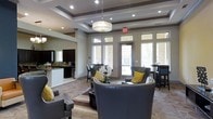 This single-floor space presents a well-appointed interior designed for comfort and functionality. The main lounge area serves as the focal point, featuring sophisticated dark leather seating with studded detailing, accented by bold yellow throw pillows that add a vibrant contrast to the neutral color palette. A prominent drum pendant light fixture hangs from a coffered ceiling with recessed lighting, lending an air of elegance to the space.

Large windows and glass-paneled doors line the exterior wall, flooding the interior with natural light while offering views of the outdoors. Transom windows above the doors further enhance the brightness of the space. The tray ceiling, framed with decorative molding and multi-level recessed lighting, adds architectural depth and dimension.

Adjacent to the lounge, a kitchenette area features dark cabinetry, a light countertop, and bar-height seating, providing a practical space for refreshments and casual dining. High-top tables and bar stools near the windows offer additional seating options. The warm-toned tile flooring ties the areas together cohesively, creating a polished, welcoming environment suited for social gatherings or relaxed activities.