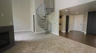 This two-story space is defined by its standout architectural centerpiece: a sleek metal spiral staircase that winds gracefully from the main level to the upper floor. The staircase, finished in brushed steel with open metal balusters, draws the eye upward and highlights the generous ceiling height of the main living area, contributing to an open, airy atmosphere. The main level is carpeted in a neutral, warm-toned broadloom that extends across the spacious living area, lending a comfortable, understated feel. The walls are finished in a soft greige tone, complementing the clean white baseboards and trim throughout. Adjacent to the living space, the flooring transitions to a darker hardwood or laminate surface, leading into a kitchen area visible in the background. The kitchen features light cabinetry and is flanked by doors providing access to additional rooms or storage. Recessed lighting illuminates the secondary spaces, while the open layout allows for flexible use of the main living zone. Overall, the space presents a clean, modern aesthetic with practical design elements suited for everyday living across both levels.