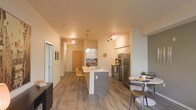 This single-floor space showcases a thoughtfully designed open-concept layout that seamlessly connects the kitchen, dining, and living areas. The kitchen serves as the centerpiece of the space, anchored by a two-tone island with a white countertop and dark wood-paneled base, complemented by bar-height seating. White upper cabinetry pairs with a dark tile backsplash, and stainless steel appliances — including a refrigerator, range, and microwave — provide a sleek, contemporary finish. Pendant lighting over the island adds focused illumination, while adjustable track lighting throughout ensures the entire space is well-lit. Warm-toned, wood-look plank flooring runs continuously across the floor, unifying the open layout and adding a welcoming feel. A compact dining area to one side accommodates a round table with mixed seating, making efficient use of the available space. The entryway, visible in the background, features a warm-toned door and clean white walls that contribute to the bright, airy ambiance. The overall design balances modern aesthetics with functional living, making it a practical and visually cohesive space.