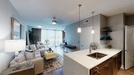 Located on E Riverside Drive in Austin, TX, this single-story unit showcases a thoughtfully designed open-concept layout that seamlessly connects the kitchen, living, and sleeping areas. The kitchen is a standout feature, centered around an expansive marble-style quartz waterfall island with an undermount sink and bar seating. Dark espresso cabinetry contrasts elegantly with the light countertops, while stainless steel appliances, a herringbone tile backsplash, and pendant lighting with track lighting overhead complete the space.

The open living area features warm hardwood-style flooring, neutral toned walls, and a ceiling fan, creating a bright and comfortable atmosphere. Floor-to-ceiling windows dressed with layered sheer and blackout curtains flood the space with natural light and provide views of the surrounding greenery. The bedroom carries the same refined aesthetic with hardwood-style flooring, large windows, and access to outdoor views.

The bathroom is clean and functional, featuring dark tile flooring, a dark wood vanity with an undermount sink, and a tub-shower combination with white subway tile surround. An in-unit laundry area with a full-size side-by-side washer and dryer, along with overhead shelving, adds everyday convenience to this well-appointed Austin residence.