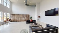 Located at 15 Abbey Ln in Danbury, Connecticut, this single-floor communal space makes an immediate impression with its dramatically high ceilings and open, airy layout. The design blends modern and industrial aesthetics seamlessly, highlighted by exposed ductwork overhead and crisp white walls that amplify the sense of volume throughout.

The main lounge area features light-toned tile flooring and is anchored by sleek black leather seating, a tufted ottoman, and a wall-mounted flat-screen television. A striking full-length reclaimed wood accent wall serves as a bold focal point, adding warmth and texture against the otherwise minimalist palette. Package lockers integrated into this wood wall provide a practical amenity within the stylish setting.

A secondary seating area offers a more relaxed atmosphere, furnished with patterned accent chairs positioned beside oversized windows that frame views of the surrounding neighborhood. These tall, multi-pane windows flood the space with abundant natural light throughout the day. A bistro-style bar counter with stools provides an additional functional area for casual use.

Light wood-look flooring transitions through the space, tying together distinct zones while maintaining a cohesive, contemporary feel. Potted tropical plants are placed throughout, softening the clean architectural lines and adding a touch of natural vitality to this well-appointed, versatile communal space.