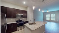 This single-floor residence offers a thoughtfully designed open-concept layout that seamlessly connects the living, dining, and kitchen areas. The kitchen is a standout feature, showcasing rich dark espresso cabinetry that extends to the ceiling for ample storage, light granite countertops with subtle speckled detailing, and a full suite of stainless steel appliances including a refrigerator, range, and built-in microwave. A tile backsplash adds a refined touch, while a peninsula with an undermount sink and pendant lighting above provides both additional prep space and a casual dining option. Light wood-style flooring runs throughout the main living areas, complementing the neutral wall tones and creating a warm, cohesive aesthetic. The living room benefits from vaulted ceilings, a ceiling fan, and large windows that invite natural light. A bedroom features sliding glass doors opening to a green outdoor view, plush carpeting, and a ceiling fan. The primary suite includes direct access to a walk-in closet and a private bathroom. The bathroom is finished with a dark vanity, granite countertop, framed mirror, and modern fixtures. Throughout the space, clean lines and consistent finishes create a polished, move-in-ready environment.