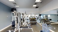 This fitness center is designed to support a full range of workout routines in a clean, functional environment. The space is outfitted with a multi-station cable machine and adjustable weight bench, making it well-suited for strength training. A tiered dumbbell rack holds an extensive set of free weights, providing options for users of all fitness levels. Along the perimeter, a row of cardio machines — including treadmills and upright bikes — offers ample opportunity for cardiovascular exercise. Large floor-to-ceiling mirrors line one wall, visually expanding the space while allowing users to monitor their form during workouts. The flooring is a durable, dark-toned material appropriate for high-traffic fitness use. Overhead, recessed panel lighting provides consistent, even illumination throughout the room, and ventilation is supported by ceiling-mounted HVAC vents. Natural light filters in through windows visible in the background, contributing to the bright and open atmosphere of the space. The overall layout is thoughtfully organized, with clear separation between the strength and cardio zones, ensuring an efficient and comfortable experience for all users.