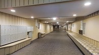 This single-floor corridor serves as a shared common area, stretching a considerable length and offering a practical, utilitarian layout. The hallway is finished with vertical wood-panel wainscoting along both walls, lending a warm, structured appearance to the space. Recessed and flush-mounted ceiling lights are evenly distributed overhead, providing consistent illumination throughout the length of the corridor.

On the left side near the entrance, a bank of individual mailboxes is mounted to the wall, accompanied by a mail drop slot and a small waste receptacle nearby. A built-in wooden bench offers a convenient seating area adjacent to the mailboxes. Informational notices and signage are posted on the walls at various points along the corridor.

The right side of the hallway features an extended upholstered bench running along the wall, providing additional seating for residents or visitors. The floor is covered in a durable, patterned carpet in neutral tones, suited for high-traffic use. The corridor's long, linear design guides occupants toward doors and additional spaces visible at the far end, emphasizing the functional nature of this well-organized communal passageway.