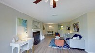 This thoughtfully designed single-floor residence offers an open-concept layout that connects the living, dining, and kitchen areas in a seamless, functional flow. The living room serves as the heart of the home, anchored by a classic white-mantled fireplace that adds warmth and character to the space. Rich hardwood-style flooring extends throughout, grounding the open layout with a sense of continuity and warmth. A ceiling fan with a dark wood finish provides both style and comfort overhead, while a textured ceiling adds subtle architectural detail. The living area is generously proportioned, accommodating a full seating arrangement alongside a dedicated workspace or desk area, making it as versatile as it is inviting. Soft, neutral wall tones in warm grey create a calm backdrop that complements the eclectic furnishings, including bold accent chairs in terracotta and navy blue tones. A patterned area rug defines the central seating zone, adding visual interest to the space. Beyond the living room, a dining area with a warm pendant light fixture offers an intimate setting for meals, while a pass-through view into the kitchen suggests a practical and connected layout. Framed artwork throughout the space adds personality and a curated aesthetic.
