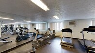 This fitness center is a fully equipped, single-floor facility designed to support a wide range of workout routines. The space features durable rubber flooring throughout, providing a safe and practical surface for exercise. Recessed fluorescent lighting illuminates the room evenly, while multiple windows along the walls allow natural light to filter in, contributing to an open and energizing atmosphere.

The cardio zone is well-appointed with multiple treadmills, an elliptical trainer, and a recumbent bike, all positioned to take advantage of the natural light from the surrounding windows. A large wall-mounted mirror on one side of the room amplifies the sense of space and allows users to monitor their form during workouts.

The strength training area includes a multi-station cable machine ideal for a variety of resistance exercises, along with a dedicated free weights section featuring a full dumbbell rack, stability balls, resistance bands, and a workout bench. A wall-mounted flat-screen television adds entertainment value to the space.

The facility is climate-controlled with wall-mounted AC units and baseboard heating, ensuring year-round comfort. The space is accessed through a glass-paneled door labeled "Fitness Center," and the overall layout is organized and practical, making efficient use of the available floor area for a diverse and functional workout environment.