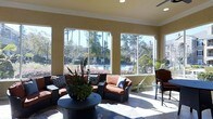 Located in Morrisville, NC, this single-floor space offers a welcoming and light-filled lounge area designed for relaxation and social gathering. The room is anchored by an expansive wall of large windows that flood the interior with abundant natural light and frame panoramic views of a sparkling outdoor pool, manicured landscaping, and mature trees beyond. Crown molding adds a refined architectural detail along the ceiling line, complementing the warm, neutral-toned walls that create a bright yet cozy atmosphere. Recessed lighting and a ceiling fan enhance comfort throughout the space. The lounge is furnished with a cohesive set of dark wicker seating — including sofas and accent chairs — accented by rich terracotta and navy cushions that add a vibrant pop of color. A round dark coffee table serves as the central gathering point, adorned with a lush tropical plant arrangement that brings a natural element indoors. A patterned area rug grounds the seating arrangement and adds visual interest to the tile flooring beneath. The overall ambiance is one of casual elegance, making this space ideal for unwinding or entertaining while enjoying the serene outdoor scenery just beyond the glass.