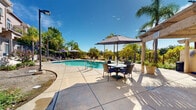 Located in Rocklin, California, this well-appointed outdoor community amenity area centers around a generously sized freeform swimming pool with clear turquoise water, offering an inviting retreat under the abundant Northern California sunshine. The pool area is complemented by a separate spa, providing a variety of aquatic options for residents and guests alike.

The surrounding concrete deck is thoughtfully designed to accommodate relaxation and social gathering. Multiple lounge chairs are positioned poolside for sunbathing, while a shaded outdoor dining area featuring a large umbrella and table-and-chair seating provides a comfortable spot for al fresco meals and conversation. An additional pergola structure adds architectural interest and further shade to the space.

Tall, swaying palm trees are planted throughout the area, enhancing the resort-like ambiance and providing natural shade and visual appeal. Lush greenery and mature trees line the perimeter fencing, creating a sense of privacy and seclusion from the surrounding landscape.

A pool house structure with a terracotta tile roof is situated along one side of the area, providing utility access and restroom facilities. The multi-story residential building beyond the pool features a Mediterranean-inspired stucco exterior with arched windows and private balconies, overlooking the pool area and contributing to the cohesive architectural character of the community.