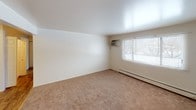 This single-floor unit offers a straightforward layout with clearly defined living, sleeping, and functional areas. The living room is carpeted in neutral tones and features a large window that allows natural light to filter in, along with a wall-mounted air conditioning unit and a baseboard heater, providing year-round climate control. An entryway door opens directly into this space, which connects openly to the kitchen.

The kitchen is efficiently arranged in a galley-style configuration, featuring dark wood cabinetry both above and below the countertops, a double stainless steel sink, laminate countertops, a gas range with an overhead ventilation hood, and a full-size refrigerator. The hardwood-style flooring in this area contrasts with the carpeted rooms and adds a practical, easy-to-clean surface.

The bedroom is a well-proportioned carpeted room with neutral white walls, a standard closet with bifold doors offering ample storage, and a window that brings in additional natural light alongside a baseboard heater. The full bathroom is compact and includes a vanity with under-sink cabinet storage, a medicine cabinet mirror above, a toilet, and a tub-shower combination with tiled surround. The overall unit presents a clean, functional interior well-suited for everyday living.