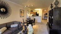 This single-floor residence offers an open-concept layout that seamlessly connects the living, dining, and kitchen areas, creating a cohesive and functional living environment. The living area features plush carpeted flooring, crown molding along the ceiling, and track lighting that provides focused illumination, lending the space a warm and inviting ambiance. A decorative arched entryway serves as an architectural accent, adding a classic touch to the interior design. The dining area is centrally positioned and flows naturally into both the living space and the kitchen, making it ideal for entertaining. The kitchen is defined by a raised breakfast bar that separates it from the main living area while maintaining visual connectivity. Dark cabinetry contrasts with lighter countertops, and the kitchen is equipped with full-size appliances. The warm, neutral color palette used throughout the space — from the walls to the flooring — creates a consistent and harmonious aesthetic. Additional details such as decorative columns and carefully placed lighting fixtures further enhance the refined character of the interior. A hallway visible from the main living area suggests additional rooms beyond, rounding out this well-appointed single-level home.