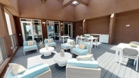 This single-story amenity space comprises two distinct functional areas that together reflect a polished, contemporary aesthetic. The outdoor terrace is an expansive, partially enclosed deck framed by warm brown walls that rise to a dramatic vaulted ceiling with skylights, allowing natural light to flood the space. The area is thoughtfully furnished with wicker lounge seating upholstered in soft aqua cushions, round white coffee tables, and a dedicated dining area with white tables and chairs. A serving station or outdoor cabinetry unit anchors one end, while glass sliding doors provide seamless access to the interior. The overall atmosphere is relaxed yet refined, suited for both casual lounging and social gatherings.

The mail room presents a sleek, modern design characterized by soft teal-painted walls, large floor-to-ceiling windows that invite abundant natural light and views of surrounding greenery, and light tile flooring. Rows of dark bronze individual mailboxes line freestanding partitions arranged in a corridor layout, creating an organized and efficient flow. Built-in package drop counters with convenient cutouts offer practical parcel handling. Framed graphic artwork adorns the walls, adding a curated, gallery-like touch to this utilitarian space.