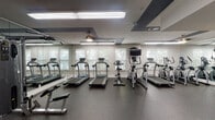 This well-equipped fitness center at 4101 Skye Ln in Cary, NC, offers a comprehensive workout environment spread across a single floor. The space is thoughtfully designed with a dark speckled rubber flooring throughout, providing both durability and safety for exercisers.

The cardio zone is lined with multiple treadmills and elliptical trainers positioned along a windowed wall with slatted blinds that allow natural light to filter in. A wall-mounted television is available in this area for entertainment during workouts. Full-length mirrors on adjacent walls enhance the sense of space and allow for form checks during exercise.

The strength training area features a multi-station cable and weight machine setup, ideal for a variety of resistance exercises. A dedicated free weights section includes a tiered dumbbell rack stocked with a range of weights and an adjustable workout bench. The functional training corner is equipped with medicine balls, resistance bands, stability balls, and other accessories stored on organized racks.

Near the entrance, a built-in bench with cubbies provides a convenient spot to store belongings, while a dual water fountain station keeps members hydrated. The ceiling features a distinctive geometric design with dark accent panels and recessed lighting, contributing to a modern, polished ambiance throughout the facility.