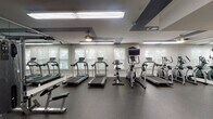 This well-equipped fitness center at 4101 Skye Ln in Cary, NC, offers a comprehensive workout environment spread across a single floor. The space is thoughtfully designed with a dark speckled rubber flooring throughout, providing both durability and safety for exercisers.

The cardio zone is lined with multiple treadmills and elliptical trainers positioned along a windowed wall with slatted blinds that allow natural light to filter in. A wall-mounted television is available in this area for entertainment during workouts. Full-length mirrors on adjacent walls enhance the sense of space and allow for form checks during exercise.

The strength training area features a multi-station cable and weight machine setup, ideal for a variety of resistance exercises. A dedicated free weights section includes a tiered dumbbell rack stocked with a range of weights and an adjustable workout bench. The functional training corner is equipped with medicine balls, resistance bands, stability balls, and other accessories stored on organized racks.

Near the entrance, a built-in bench with cubbies provides a convenient spot to store belongings, while a dual water fountain station keeps members hydrated. The ceiling features a distinctive geometric design with dark accent panels and recessed lighting, contributing to a modern, polished ambiance throughout the facility.