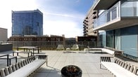 This outdoor rooftop terrace offers a well-appointed urban retreat with sweeping views of the surrounding cityscape. The space is finished with large-format tiled pavers that provide a clean, modern aesthetic underfoot, while a glass-panel railing system along the perimeter ensures unobstructed sightlines across the skyline.

A central fire pit filled with decorative stones serves as a natural focal point, making the terrace functional across multiple seasons. Modern white lounge chairs and benches are thoughtfully arranged throughout the space, providing multiple seating configurations suited for both casual relaxation and social gatherings. Additional bench-style seating is positioned along the edges of the terrace, taking advantage of the panoramic views.

The terrace is bordered by a mix of residential high-rises and lower brick buildings, offering a layered urban backdrop. Overhead, the open sky and ample natural light enhance the airy atmosphere of the space. The combination of contemporary furnishings, durable materials, and a well-considered layout make this rooftop terrace a versatile and inviting outdoor amenity.