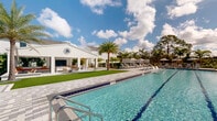 Located in Bonita Springs, Florida, this outdoor amenity facility offers a resort-style experience designed for both recreation and relaxation. The centerpiece is a generously sized lap pool with clearly marked swim lanes, framed by a wide, elegantly paved pool deck finished in light-toned stone pavers. The pool features a wading shelf along one end, ideal for lounging in shallow water, and is surrounded by an array of chaise lounges and shaded cabanas with striped umbrellas.

Adjacent to the pool is a covered outdoor pavilion, architecturally styled with white-painted walls, a gabled roofline, and a standing seam metal accent, echoing the coastal contemporary design of the surrounding community. The pavilion is equipped with a large outdoor screen and comfortable seating areas, making it a versatile gathering space for residents. A vibrant green turf lawn connects the pavilion to the pool deck, adding a fresh, manicured aesthetic.

Tall palm trees and mature tropical plantings frame the entire area, providing natural shade and privacy while contributing to the lush Florida ambiance. The facility is bathed in abundant natural sunlight, with open sky views that highlight the bright, airy character of this outdoor space. The overall design reflects a polished, community-focused lifestyle destination.