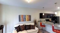 This well-appointed single-floor space offers a seamlessly connected living and kitchen area designed for both comfort and function. The living area is anchored by a cream-colored sofa accented with richly patterned dark throw pillows, complemented by a bold, colorful abstract wall piece that adds a vibrant focal point to the otherwise neutral palette of soft gray walls and light flooring. Track lighting overhead provides bright, directed illumination throughout the space. Transitioning into the kitchen area, dark espresso cabinetry creates a striking contrast against granite-style countertops, lending a sleek and contemporary aesthetic. A peninsula counter with a built-in bar overhang is paired with a modern red bar stool, offering a casual dining or entertaining option. A pendant light hangs above the counter, adding a warm, inviting glow to the kitchen zone. Decorative dried branches in a floor vase bridge the two areas with an organic, natural touch. The overall ambiance is modern and cohesive, with thoughtful design details that balance functionality with style throughout this compact yet efficiently arranged single-level layout.