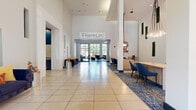 Franklin at Samuels Ave presents a thoughtfully designed communal space that blends contemporary style with welcoming functionality. The grand lobby serves as the centerpiece of the community, boasting soaring ceilings, large-format tile flooring, and an abundance of natural light streaming through floor-to-ceiling glass entry doors. The space is anchored by the prominently displayed Franklin at Samuels Ave signage and accented with living moss wall panels, vibrant artwork, and elegant pendant lighting that add distinct character throughout.

A dedicated lounge area with plush navy blue seating and a patterned area rug offers residents a comfortable space to relax or socialize, while a sleek reception desk with modern blue chairs provides a polished point of service. The amenity kitchen features a striking marble-topped island with leather barstools, mosaic tile backsplash, stainless steel appliances, and navy cabinetry. An adjacent dining area with a rustic wood table and leather chairs creates an inviting setting for casual meals. A separate lounge room with dark hardwood floors, a white stacked-stone fireplace, curated artwork, and lush greenery adds a warm, refined ambiance to round out the communal offerings.