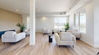 Located at 1650 Ferry Wharf Road in Kelowna, BC, this single-floor amenity space offers a well-appointed, modern environment designed for both collaborative work and relaxed gathering. The layout is divided into two distinct functional zones that together create a versatile and inviting atmosphere throughout.

The conference area features a large rectangular table surrounded by ergonomic black office chairs, complemented by a full-size wall-mounted display screen set against a warm slatted wood accent wall. A frosted window provides natural light while maintaining privacy, and light hardwood-style flooring ties the room together with a clean, professional aesthetic.

The adjoining lounge area is bright and open, bathed in natural light from expansive windows that frame sweeping outdoor views of the surrounding Kelowna landscape. Plush, curved cream armchairs are arranged around sleek black side tables and small round café-style tables, creating an inviting setting for informal meetings or relaxation. Touches of greenery from potted palms add warmth to the otherwise crisp, neutral palette of white walls and light flooring.

A wood slat accent wall anchors the lounge's seating area, echoing the design language of the conference room and lending visual cohesion to the space. Recessed lighting throughout ensures a consistently well-lit environment, making this a functional and stylish space suited to a variety of uses.
