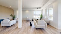 Located at 1650 Ferry Wharf Road in Kelowna, BC, this single-floor amenity space offers a well-appointed, modern environment designed for both collaborative work and relaxed gathering. The layout is divided into two distinct functional zones that together create a versatile and inviting atmosphere throughout.

The conference area features a large rectangular table surrounded by ergonomic black office chairs, complemented by a full-size wall-mounted display screen set against a warm slatted wood accent wall. A frosted window provides natural light while maintaining privacy, and light hardwood-style flooring ties the room together with a clean, professional aesthetic.

The adjoining lounge area is bright and open, bathed in natural light from expansive windows that frame sweeping outdoor views of the surrounding Kelowna landscape. Plush, curved cream armchairs are arranged around sleek black side tables and small round café-style tables, creating an inviting setting for informal meetings or relaxation. Touches of greenery from potted palms add warmth to the otherwise crisp, neutral palette of white walls and light flooring.

A wood slat accent wall anchors the lounge's seating area, echoing the design language of the conference room and lending visual cohesion to the space. Recessed lighting throughout ensures a consistently well-lit environment, making this a functional and stylish space suited to a variety of uses.
