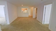 This single-floor residence offers a clean, functional layout designed for comfortable everyday living. The main living area is spacious and open, finished with wall-to-wall beige carpeting that adds warmth and a neutral palette throughout. Smooth white walls and recessed lighting contribute to a bright, well-lit atmosphere without relying solely on natural light.

A defining feature of the space is the pass-through kitchen window, which connects the kitchen to the main living area and promotes an open, connected feel. The kitchen is outfitted with warm oak-toned cabinetry and includes a full-size white refrigerator, providing practical storage and appliance space. The kitchen flooring transitions to a hard surface, distinguishing it from the carpeted living area.

From the living room, a doorway leads to additional rooms beyond, suggesting a thoughtful separation of living and private spaces. The six-panel interior doors are a consistent design element throughout, lending a classic, traditional aesthetic to the unit. The overall ambiance is calm and neutral, offering a versatile blank canvas well-suited for a variety of furnishing styles and personal touches.
