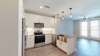 This single-floor space presents a thoughtfully designed, contemporary interior with a cohesive neutral color palette throughout. The kitchen serves as a standout feature, equipped with crisp white shaker-style cabinetry, a light stone tile backsplash, and a full suite of stainless steel appliances including a French door refrigerator, electric range, built-in microwave, and dishwasher. A peninsula with a gray quartz countertop and an integrated stainless steel farmhouse sink anchors the kitchen, illuminated by a pair of geometric pendant lights that add a modern industrial touch. Under-cabinet lighting enhances the workspace.

The open-concept layout connects the kitchen seamlessly to the living area, which features wood-look plank flooring and neutral warm-toned walls. A set of French doors with blinds opens to an outdoor space, allowing natural light to fill the room, complemented by a ceiling fan overhead.

The bedroom is finished with plush carpet, a ceiling fan, and a window with blinds that allows for natural light. The bathroom is bright and clean, featuring large-format tile flooring, a glass-enclosed walk-in shower with tile surround, a white vanity with an undermount sink, and a well-lit mirror.