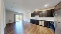 Located at 77 S Adams St in Oswego, IL, this well-appointed one-bedroom, one-bathroom unit offers a thoughtfully designed single-level layout. The kitchen is a standout feature, showcasing rich dark cabinetry with ample upper and lower storage, crisp white countertops, a stainless steel appliance suite including a gas range, built-in microwave, dishwasher, and refrigerator, and light hardwood-style flooring that flows throughout the unit. The open sightlines from the entry into the kitchen create a welcoming, functional space. The bedroom is comfortably sized, offering a window with natural light and views of the surrounding greenery, along with a dedicated walk-in closet equipped with built-in shelving and hanging rods. The bathroom continues the cohesive design language with dark cabinetry, a white countertop vanity, a large mirror, and clean tile-style flooring. A private balcony provides serene outdoor views of open green lawns and mature trees, set against the backdrop of the well-maintained multi-story building exterior with classic brick and craftsman-style architectural details. Clean white walls and consistent flooring throughout give the space a bright, modern ambiance.
