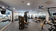 Located at 169 Johnson Rd in Morris Plains, NJ, this well-appointed fitness center offers a comprehensive exercise facility across a single, generously sized floor. The space is thoughtfully designed to accommodate a wide range of workout styles, blending modern aesthetics with functional equipment.

The main exercise area features durable rubber flooring throughout the strength and cardio zones, housing a robust selection of equipment including multi-station cable machines, adjustable weight benches, a full dumbbell rack, and a row of cardio machines such as ellipticals and upright bikes. Large wall-mounted mirrors expand the sense of space and allow for form monitoring during workouts. Recessed lighting and ceiling fans ensure the area remains bright and comfortable.

A distinct cycling studio is separated by sleek floor-to-ceiling glass partitions with black framing, creating a dedicated spin area with light wood flooring, indoor cycling bikes, and a decorative sculptural wall accent that lends an upscale ambiance. A wall-mounted flat-screen TV is also present for entertainment. The open layout, modern finishes, and carefully curated equipment selection make this fitness center a well-rounded facility suited for strength training, cardio, and group cycling workouts alike.