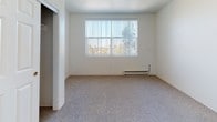 This single-floor unit offers a clean, straightforward layout designed for comfortable everyday living. The kitchen is efficiently arranged in a U-shaped configuration, featuring light-toned cabinetry with ample storage, granite-look countertops, white appliances including a refrigerator and range, and tile flooring that adds easy-clean practicality to the space. A pass-through opening connects the kitchen to the adjoining living area, creating a sense of openness between the two spaces.

The living and bedroom areas are finished with wall-to-wall carpeting in a neutral tone, providing warmth underfoot. Large windows dressed with vertical blinds allow generous natural light to filter in while offering views of surrounding greenery. Baseboard heating units are present in the carpeted rooms, ensuring consistent warmth. A pendant light fixture in one of the rooms adds a simple decorative touch.

The bathroom is designed with accessibility in mind, featuring stainless steel grab bars near the toilet and along the wall, a wall-mounted sink beneath a vanity mirror with overhead lighting, and patterned tile flooring. A frosted window provides natural light while maintaining privacy. The overall palette throughout the unit is light and neutral, contributing to a bright, open ambiance suitable for a range of occupants.