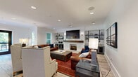 Located at 1030 Jack Primus Rd in Charleston, SC, this well-appointed single-floor space offers a generous and thoughtfully designed layout encompassing multiple living, dining, and kitchen areas connected by a central hallway. The interior design masterfully blends industrial and transitional styles, featuring rich dark wood beam accents, bold black steel-framed glass partition walls, and light wood-look flooring throughout. The main living area is anchored by a striking white brick fireplace with a reclaimed wood mantle, a wall-mounted display, and flanking industrial pipe shelving units — all bathed in natural light from large traditional windows. A second lounge area provides a more intimate setting with plush leather seating, sheer floor-to-ceiling drapery, and direct access to an outdoor deck surrounded by lush greenery. The kitchen is equipped with stainless steel appliances, gray subway tile backsplash, slate-toned cabinetry, and distinctive patterned tile flooring. A combined dining and living area features a built-in bar counter with wood barstools, pendant lighting, and an open sightline to adjacent spaces. The dining nook offers a rustic wood table beside natural-light-filled windows with sheer draping. Throughout, the space maintains a cohesive, sophisticated ambiance ideal for gathering and community use.