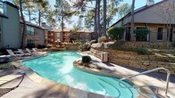 This single-story unit is situated within a well-established residential community characterized by mature pine trees, multi-story buildings with brick and wood exteriors, and carefully maintained landscaping. The outdoor common area serves as a focal point of the community, centering on a generously sized freeform swimming pool with clear turquoise water. The pool is surrounded by a stone-tile deck and bordered by tiered stacked-stone retaining walls that incorporate planted beds with shrubs and ornamental grasses, creating a naturalistic, layered environment. Lounge chairs are positioned along the poolside, and the towering pines provide natural shade throughout the day.

The interior of the unit features dark wood-style plank flooring that runs continuously through the main living areas, lending a warm, cohesive feel. The living room is open and comfortably scaled, appointed with a ceiling fan and textured ceiling. Neutral white walls keep the space bright and versatile. A hallway with large windows fitted with horizontal blinds allows natural light to filter through while maintaining privacy. Recessed lighting and an exit-lit corridor reinforce the practical, well-maintained character of the interior layout throughout.