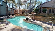 This single-story unit is situated within a well-established residential community characterized by mature pine trees, multi-story buildings with brick and wood exteriors, and carefully maintained landscaping. The outdoor common area serves as a focal point of the community, centering on a generously sized freeform swimming pool with clear turquoise water. The pool is surrounded by a stone-tile deck and bordered by tiered stacked-stone retaining walls that incorporate planted beds with shrubs and ornamental grasses, creating a naturalistic, layered environment. Lounge chairs are positioned along the poolside, and the towering pines provide natural shade throughout the day.

The interior of the unit features dark wood-style plank flooring that runs continuously through the main living areas, lending a warm, cohesive feel. The living room is open and comfortably scaled, appointed with a ceiling fan and textured ceiling. Neutral white walls keep the space bright and versatile. A hallway with large windows fitted with horizontal blinds allows natural light to filter through while maintaining privacy. Recessed lighting and an exit-lit corridor reinforce the practical, well-maintained character of the interior layout throughout.
