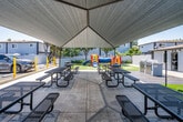Located at 811 S Sullivan St in Santa Ana, California, this outdoor community amenity space is designed to provide residents with functional recreational areas in a well-maintained setting. The centerpiece of the space is a large shade structure supported by a striking triangular tensile canopy, beneath which several metal picnic tables with bench seating are arranged on a concrete pad — ideal for outdoor gatherings and communal dining. Adjacent to the covered seating area, a charcoal grill provides additional convenience for outdoor cooking. The playground area is a standout feature, equipped with a vibrant, multi-component play structure that includes slides, tunnels, and climbing elements, all set on lush artificial turf that ensures a clean and safe surface for children. Mature trees are scattered throughout the grounds, offering natural shade and contributing to the inviting atmosphere. A perimeter block wall provides clear boundaries and a sense of privacy for the space. Pathways connect the various zones, including access to on-site parking and adjacent buildings. Informational signage is posted near the entrance, and the overall grounds are kept clean and well-organized. The open layout and Southern California sunshine make this a bright, welcoming community hub year-round.