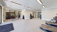 Located at 3450 Miller Dr in Chamblee, Georgia, this well-appointed fitness center offers a thoughtfully designed, single-floor layout that maximizes functionality and comfort. The expansive exercise room is the heart of the facility, featuring plush carpet flooring, crown molding, and bright recessed linear lighting that creates a clean, energizing atmosphere. The cardio section is equipped with multiple treadmills, an elliptical, and a stationary bike, all positioned near large floor-to-ceiling glass doors that flood the space with natural light and provide direct views of — and access to — an outdoor pool area. The strength and training zone houses a punching bag, weight bench, dumbbell rack, resistance machines, and various functional fitness accessories mounted on a dedicated wall rack, offering a comprehensive workout experience. Two restrooms serve the facility, each finished with light hardwood-style flooring, neutral gray walls, crown molding, and modern fixtures. The women's restroom includes an ADA-accessible design with a wall-mounted sink and paper towel dispenser. A tastefully decorated secondary restroom adds a refined touch with abstract wall art and a stylish wood-and-charcoal storage cabinet. Connecting hallways provide smooth circulation throughout the space, contributing to an overall ambiance that is clean, modern, and welcoming.