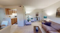 This thoughtfully designed 1-bedroom, 1-bathroom residence offers a complete and functional layout across a single floor. The open living room is a welcoming space featuring soft carpeting, neutral walls, and ample room for seating, centered around a wooden coffee table and accented with decorative artwork and warm lighting. Large windows with decorative curtains allow natural light to filter in, creating a bright and comfortable atmosphere.

Adjacent to the living room, the dining area accommodates a glass-top dining table with upholstered chairs, finished with a chandelier overhead and a window view to the outdoors. The galley-style kitchen is efficiently designed with warm wood cabinetry lining both walls, white countertops, a gas range, a full-size refrigerator, and a double sink — maximizing every inch of available space.

The bedroom is a comfortable retreat featuring plush carpeting, a large window with outdoor views, roller shades, and curtain panels for privacy. The full bathroom includes a tiled shower-tub combination, chrome fixtures, and a clean, classic finish with white tile flooring. A dedicated closet and hallway complete the layout, contributing to practical day-to-day living in a compact yet complete residence.