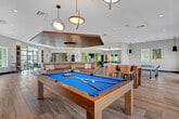 Located in Corona, California, this thoughtfully designed two-floor amenity facility offers a generous and versatile gathering space ideal for socializing and recreation. The main floor is anchored by a sophisticated clubroom featuring warm wood-slatted ceiling accents, a striking built-in sectional sofa in rich cognac leather, and light wood-look tile flooring throughout. Dramatic arched black-framed windows flood the interior with natural light while framing views of the surrounding community. A billiards area showcases a vibrant blue pool table beneath a sculptural pendant light, with decorative geometric patterned wall panels adding visual depth. Adjacent game spaces include a dedicated cornhole and recreation room finished with turf flooring, offering a casual, playful atmosphere. The upper level opens to a semi-covered outdoor terrace with ceiling fans and sweeping views of the community pool and neighboring rooftops. A fully outfitted outdoor kitchen and bar area features a white quartz waterfall-edge counter with dark brick tile detailing, a wall-mounted TV set against an ornate patterned tile backdrop, and pendant lighting. Arched openings frame the terrace lounge area, which is furnished with woven rattan seating and colorful artwork, completing this well-appointed community amenity space.