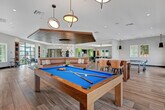 Located in Corona, California, this thoughtfully designed two-floor amenity facility offers a generous and versatile gathering space ideal for socializing and recreation. The main floor is anchored by a sophisticated clubroom featuring warm wood-slatted ceiling accents, a striking built-in sectional sofa in rich cognac leather, and light wood-look tile flooring throughout. Dramatic arched black-framed windows flood the interior with natural light while framing views of the surrounding community. A billiards area showcases a vibrant blue pool table beneath a sculptural pendant light, with decorative geometric patterned wall panels adding visual depth. Adjacent game spaces include a dedicated cornhole and recreation room finished with turf flooring, offering a casual, playful atmosphere. The upper level opens to a semi-covered outdoor terrace with ceiling fans and sweeping views of the community pool and neighboring rooftops. A fully outfitted outdoor kitchen and bar area features a white quartz waterfall-edge counter with dark brick tile detailing, a wall-mounted TV set against an ornate patterned tile backdrop, and pendant lighting. Arched openings frame the terrace lounge area, which is furnished with woven rattan seating and colorful artwork, completing this well-appointed community amenity space.