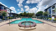 This outdoor amenity space is a well-appointed resort-style retreat centered around a generously sized swimming pool with striking turquoise water. The pool features a curved, freeform design with brick-trimmed edges and is complemented by an attached circular spa/hot tub, also finished with classic brick detailing. A concrete pool deck surrounds the water, providing ample room for lounge chairs and outdoor seating arrangements positioned to take full advantage of the open sky above.

The space is framed by multiple pergola structures that offer shaded gathering areas with outdoor furniture, ideal for relaxing or socializing. A central landscaped walkway lined with ornamental grasses, yucca plants, and decorative gravel leads from the entrance toward the pool, creating a welcoming approach flanked by towering sabal palm trees and flowering crape myrtles that add vibrant seasonal color.

The surrounding landscape is thoughtfully designed, incorporating layered plantings of tropical and subtropical species that lend the area a lush, resort-like ambiance. A multi-story residential building with balconies provides a backdrop to the pool deck, and perimeter fencing defines the boundaries of the space. The overall design prioritizes both visual appeal and functional outdoor living, making it a standout amenity for residents.