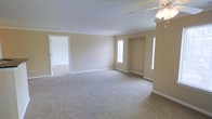 This single-floor space presents a spacious, open living area finished with wall-to-wall carpeting in a light neutral tone, complementing the warm beige walls and crisp white trim throughout. Crown molding runs along the ceiling perimeter, adding a subtle architectural detail that elevates the overall finish of the room. A ceiling fan is mounted overhead, providing both functional air circulation and a central lighting element. Natural light enters through multiple windows, including a pair of narrow vertical windows that frame a recessed niche on one wall, creating a distinctive architectural feature and adding visual depth to the room. An additional window on the adjacent wall further brightens the interior. White baseboards provide clean contrast against the neutral walls. A doorway on one side opens to a secondary room, also carpeted and finished in the same light palette, suggesting a consistent design language throughout the space. The overall ambiance is calm and understated, with a move-in-ready feel suited to a variety of uses and furnishing styles.