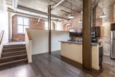 This industrial-style loft showcases the character of a thoughtfully converted historic building, blending raw architectural elements with modern finishes throughout. The open-plan living and kitchen area is anchored by striking exposed brick walls, concrete ceilings with visible steel ductwork, and bold structural columns that speak to the building's industrial heritage. Dark hardwood floors run through the main living space, complementing the rugged aesthetic.

The kitchen is a standout feature, offering dark cabinetry, granite countertops, a central island with additional counter seating, and stainless steel appliances including a refrigerator, range, and microwave. Vintage-style pendant lights hang above, enhancing the loft's industrial charm. A staircase leads to an upper landing area, adding a sense of vertical dimension to the layout.

The bedroom features original exposed brick walls and large windows that invite generous natural light, with plush carpeting providing a warm contrast to the raw surroundings. Two full bathrooms are finished in clean, neutral tones with tub/shower combinations and tile flooring. Together, these elements create a space that is both historically rich and comfortably livable.