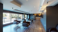 Located at 1507 Gordon St in Los Angeles, CA, this single-floor lounge space presents a sleek, contemporary design throughout. The main lounge area is anchored by large floor-to-ceiling windows fitted with roller shades, allowing for adjustable natural light and views of the surrounding streetscape. Structural concrete columns add an industrial character that complements the modern aesthetic.

The seating area is thoughtfully arranged with dark leather club chairs and accent tables, creating multiple intimate conversation zones across the wood-plank flooring. Deep blue-gray accent walls provide a bold, cohesive backdrop throughout the space, while recessed lighting and wall-mounted sconces ensure warm, even illumination.

A dedicated media nook features a wall-mounted flat-screen television flanked by accent lighting and additional lounge seating, offering a comfortable retreat for viewing. The kitchenette area is well-equipped with warm wood cabinetry, a stainless steel refrigerator, a sink with countertop workspace, and small appliances including a coffee maker and microwave — making the space fully functional for extended gatherings. The open layout and cohesive design language create a welcoming, professional ambiance suited for both casual socializing and community use.